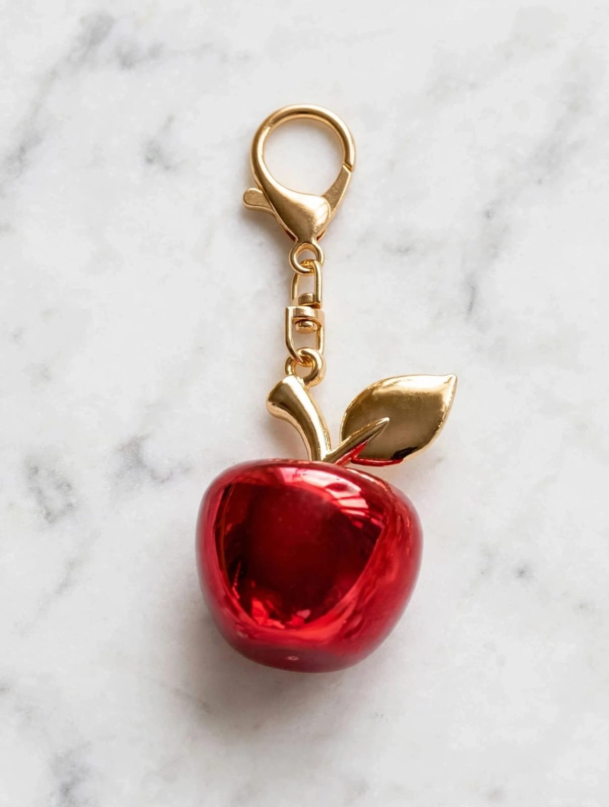 Glossy red apple keychain charm with gold leaf and lobster clasp on a white marble surface
